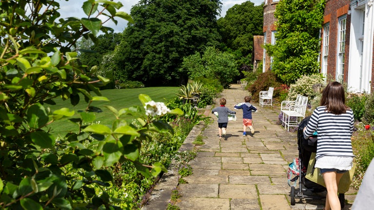 A paved garden path runs alongside a brick house with white doors and windows, bordered by lush plants and flowers. Two children run ahead along the path while an adult pushes a stroller behind them, with a large green lawn and tall trees stretching out to the left in bright summer sunshine.